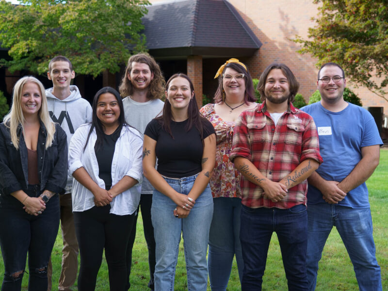 A group of 8 people standing on green grass in front of a brick building