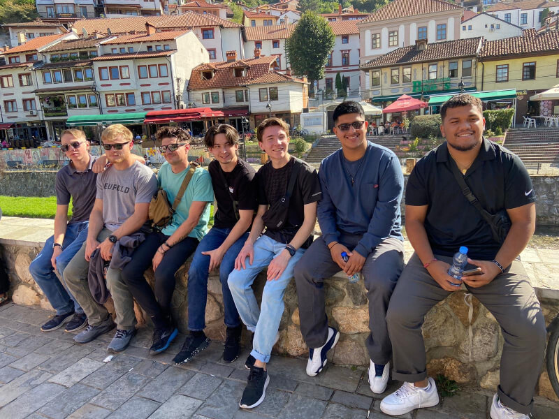 A group of guys sitting on a low stone wall in front of several buildings